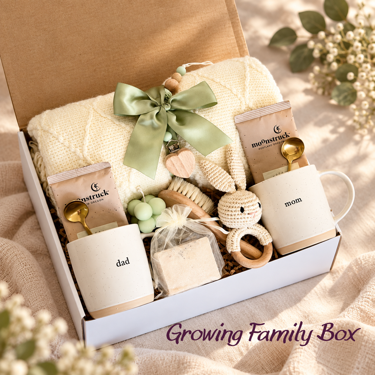Growing Family new parents gift basket in a box with mom and dad mugs, cream knit blanket, gold-colored spoons, packaged items, and bunny ring accessory.